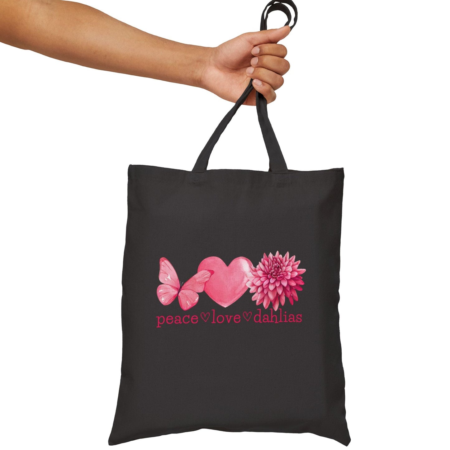 Black tote bag with pink heart, butterfly, and flower design held by a hand on a white background