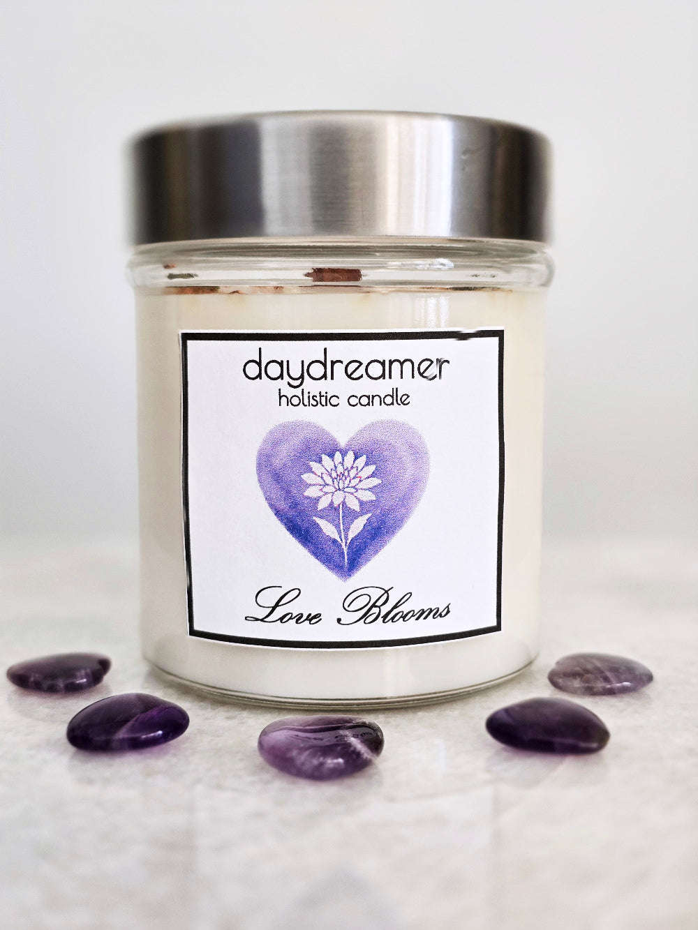 Candle jar labeled 'daydreamer holistic candle Love Blooms' with a purple heart and white dahlia flower on a white background.