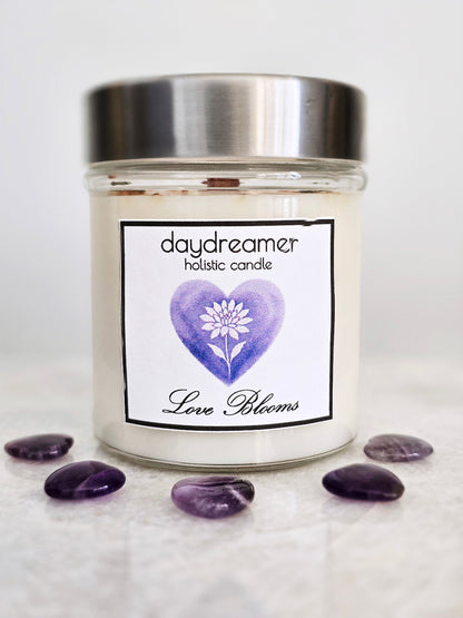 Candle jar labeled 'daydreamer holistic candle Love Blooms' with a purple heart and white dahlia flower on a white background.